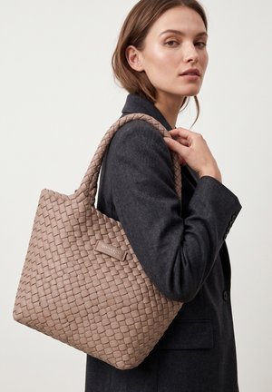Woman in dark blazer carrying large taupe woven handbag over shoulder against plain background.