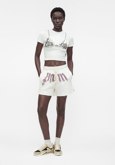 Γυναίκα που φοράει λευκό κοντό t-shirt με εκτύπωση σουτιέν, λευκά σόρτς με σκοινί και γραφικό κείμενο, κάλτσες και μπεζ-μαύρα αθλητικά παπούτσια, στέκεται με τα χέρια στις τσέπες.