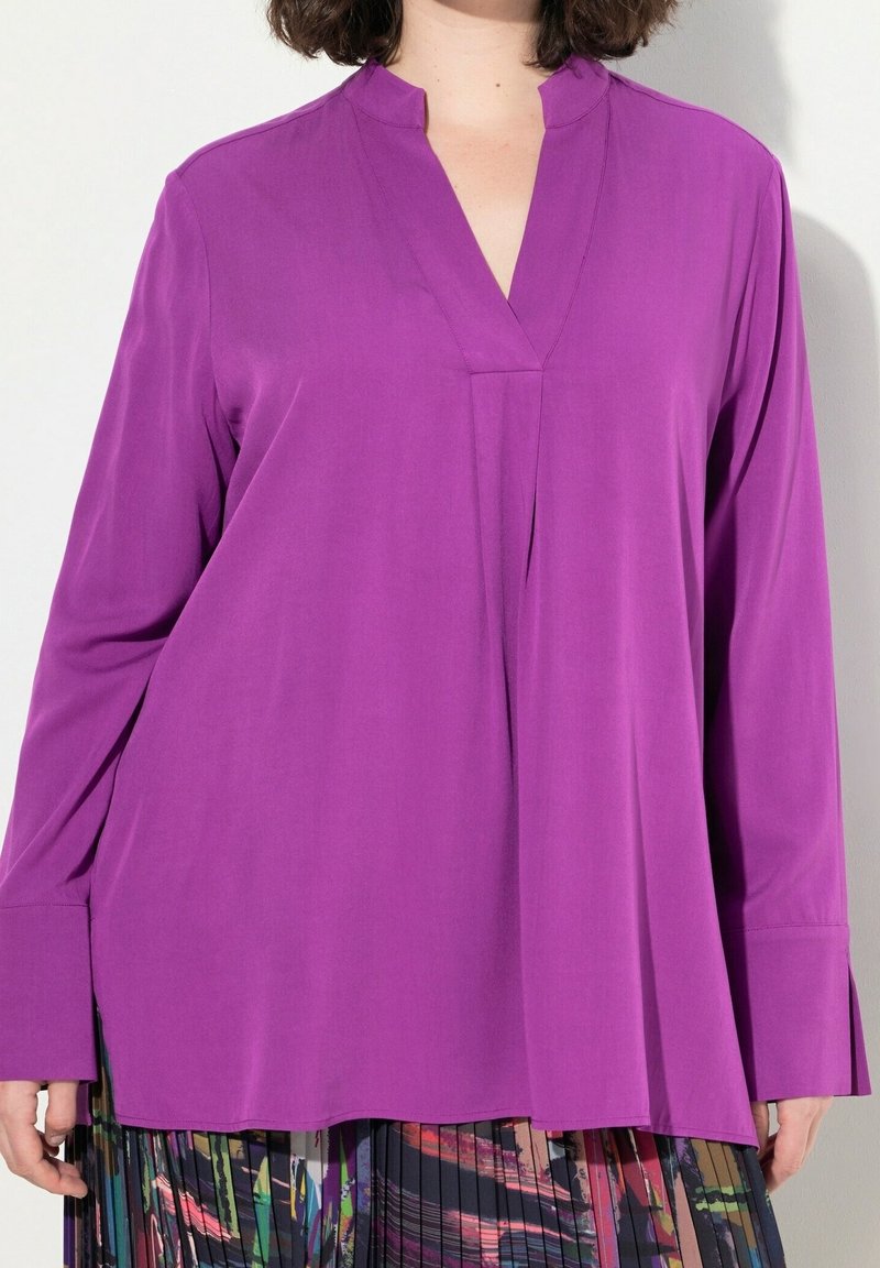 Femme portant un chemisier violet à manches longues avec un col en V et une jupe plissée à motifs sur un fond clair uni.