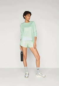 Lichtgroene lange mouwen sporttop in combinatie met bijpassende shorts. Beschermt een losse fit en vochtafvoerende stof. Grijze sneakers en een waterfles.