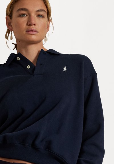 Pulover polo bleu marin din material moale, cu guler, două butoane și un logo brodat pe partea stângă a pieptului.