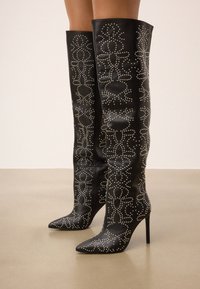 Botas de cuero negro sobre la rodilla con punteras afiladas, adornadas con tachuelas plateadas en un patrón floral, con tacones de estilete.