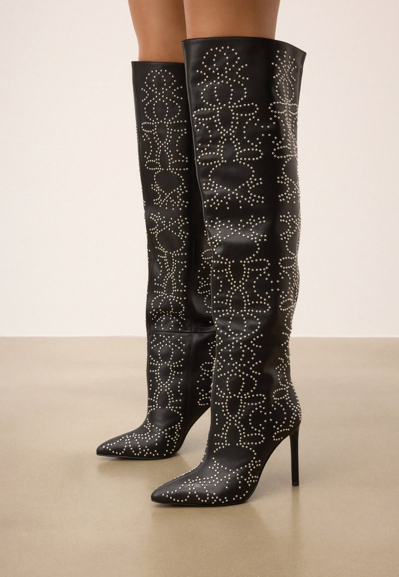 Botas de cuero negro sobre la rodilla con punteras afiladas, adornadas con tachuelas plateadas en un patrón floral, con tacones de estilete.