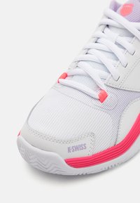 Hvit idrettssko med mesh-overdel, rosa detaljer og subtile grå striper; med sentral snøring og teksturert såle. Merkevaren er synlig.