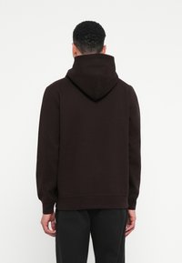 Muž s krátkymi kučeravými vlasmi, oblečený v tmavohnedej mikine a čiernych nohaviciach, stojí chrbtom k jednému bielemu pozadiu.