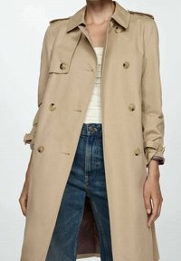 Beige trench coat with double-breasted button closure, shoulder epaulettes, and adjustable cuffs. Paired with light top and denim jeans.