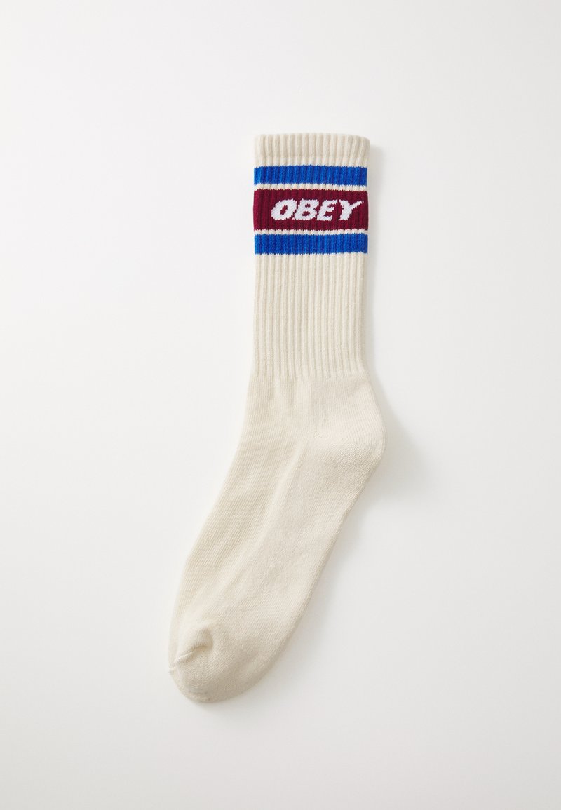 Krémová ponožka s rebrovanou textúrou, s červeným, modrým a bielym logom s nápisom "OBEY" na vrchu.