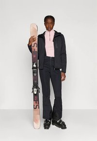 Vrouw in zwarte skibroek, zwarte jas en roze top, die een zalmkleurige ski met zwarte bindingen vasthoudt, met zwarte ski-laarzen op een grijze vloer.