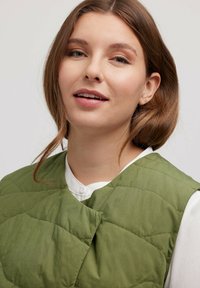 Gestepptes olivgrünes Weste mit einem einfachen runden Halsausschnitt und Knopfverschluss, über einem weißen Hemd mit Knöpfen getragen. Weiche Textur mit sichtbaren Nähten.