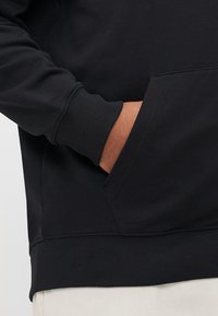Crna majica s kapuljačom koja ima kenguru džep, rebraste manžete i rub, izrađena od mekog, teksturiranog materijala. Bez vidljivih logotipa ili uzoraka.