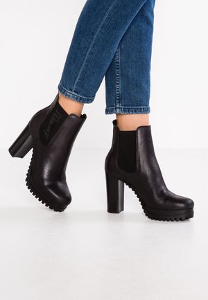 Person, die dunkelblaue Jeans und schwarze, hochhackige Stiefeletten mit klobigen Sohlen vor weißem Hintergrund trägt.