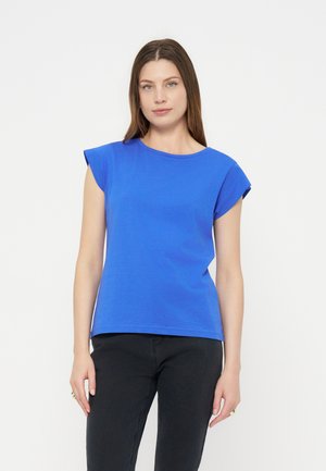 Femeie cu păr brun drept, purtând un top albastru luminos fără mâneci și pantaloni negri, stând în fața unui fundal alb simplu.