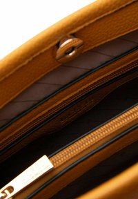 Senfgelbes Lederhandtasche mit strukturierter Oberfläche, ausgestattet mit einer Reißverschlusstasche und einem glatten Futter. Beinhaltet ein Metallschild mit Markenlogo.
