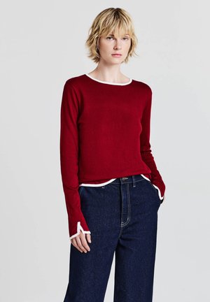 Rotes Langarmoberteil mit einem weißen Rundhalsausschnitt und Bündchen, mit einer leicht verkürzten Passform, kombiniert mit dunkelblauen hochgeschnittenen Jeans.