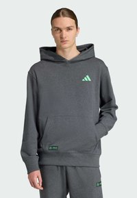 Siva dukserica s kapuljačom i kenguruyang džepom te zelenim Adidas logom. Mekana tkanina, podesiva kapuljača, rebrasti rukavi i porub; uparena s odgovarajućim hlačama.