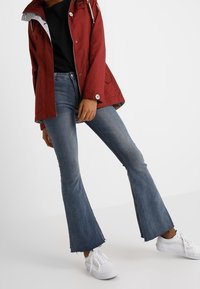 Giacca rossa con cappuccio, bottoni e cordini; t-shirt nera; jeans blu a campana con orlo sfilacciato; sneakers bianche.