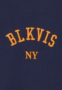 Tengerészkék szövet narancssárga hímzett „BLKVIS NY” felirattal, ívelt dizájnnal. Simább textúra, mintás díszítés nélkül.