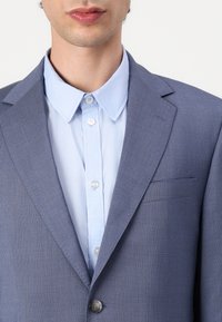 Modra prugasta blazer z ožjimi reverji, žepom na prsih in enojnim gumbom, oblečen preko svetlo modre srajce s kragnico.