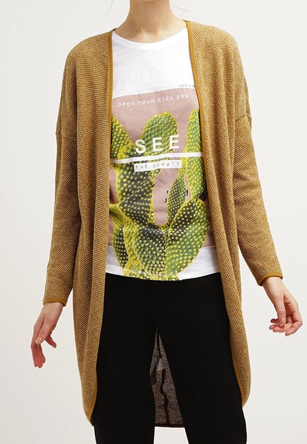 Un cardigan en maille beige porté par-dessus un t-shirt graphique blanc avec un motif de cactus. Un pantalon noir est porté en dessous, mettant en valeur la tenue superposée.