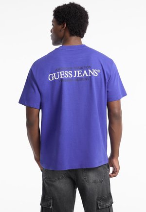 Ljubičasta pamučna majica s kratkim rukavima, s "GUESS JEANS" logom na leđima, opuštenog kroja, u kombinaciji s tamnim cargo hlačama.