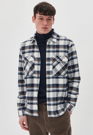 Plaid flannel shirt in white, navy, and brown; features two chest pockets, button-up front, and long sleeves. Worn over a dark turtleneck.