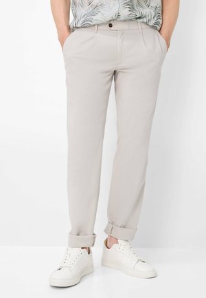 Uomo che indossa pantaloni beige chiaro risvoltati, sneakers bianche e una camicia a maniche corte con motivo a foglie, con le mani in tasca.