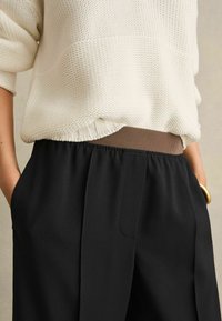 Pullover lavorato a maglia color crema con scollatura tonda, abbinato a pantaloni neri con una vita marrone. I pantaloni hanno tasche laterali e una texture liscia.