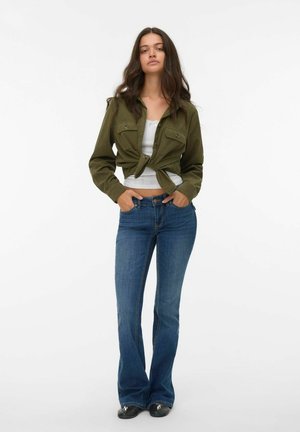 Olijfgroene cropped jack met twee borstzakken, gecombineerd met een lichtgrijze tanktop en flared blauwe jeans. Schoenen met metalen accenten.