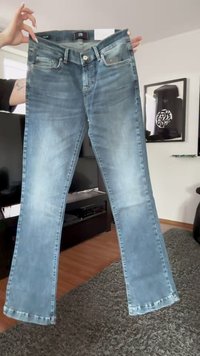 Calças de ganga de cor azul clara com um design de perna reta, bolsos frontais, fecho com botão e textura desbotada. Etiqueta visível na cintura.