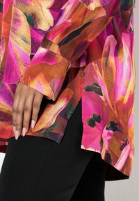 Hånd hvilende på hoften iført langærmet skjorte med levende pink, orange og gul blomstermønster, kombineret med sorte bukser.