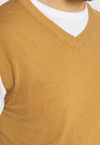 V-hals, ærmeløs sweater i gyldenbrun strik med fin ribkant ved halsudskæringen og kanten, båret over en hvid t-shirt med rund hals.