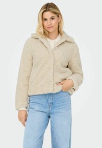Beige fuzzy jacket with a collar, featuring a button closure and ribbed cuffs, paired with light blue high-waisted jeans.