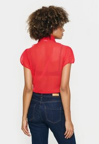 Rote transparente Bluse mit puffigen Ärmeln und einem Rüschenausschnitt, kombiniert mit dunkelblauen Jeans aus Denim mit strukturierten Taschen.