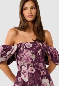 Vrouw in een paarse off-shoulder jurk met grote roze en witte bloempatronen, die iets opzij kijkt met los bruin haar.
