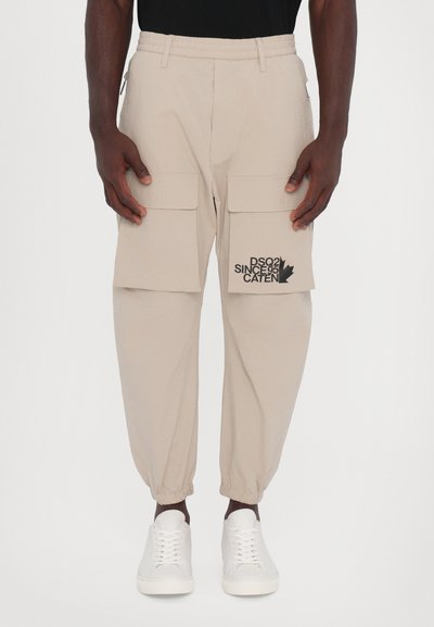 Pantalons cargo beiges avec une taille élastique, deux poches cargo à l'avant et un logo graphique noir sur la cuisse gauche. Chevilles retroussées.
