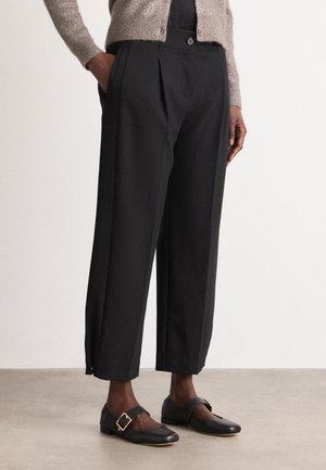 Black pleated trousers made of smooth fabric, featuring a relaxed fit and side pockets. Worn with black shoes that have a buckle detail.