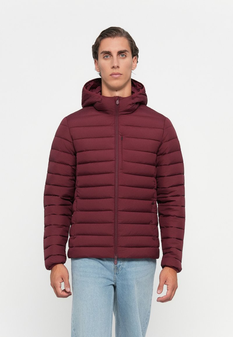 Ung mand iført en bordeaux farvet, quilted hættetrøje med lynlås foran og lyseblå jeans, stående mod en ensfarvet hvid baggrund.