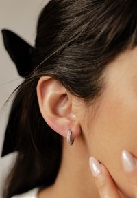 Boucle d'oreille en argent avec une surface texturée et de petites accents en pierre, présentée sur l'oreille d'un modèle ayant de longs cheveux noirs et des ongles manucurés.