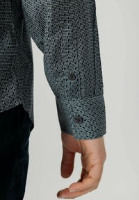 Camicetta a maniche lunghe di colore verde scuro con un motivo geometrico di linee bianche. Presenta bottoni neri ai polsi e un orlo leggermente curvato.