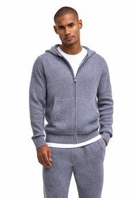 Grauer, gerippter Kapuzenpullover mit Reißverschluss, verstellbarer Kapuze, Vordertaschen und Strickstruktur, getragen über einem weißen T-Shirt.