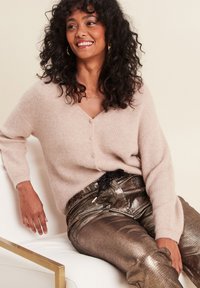 Lichtroze V-hals sweater van zacht, geribbeld materiaal met knopen aan de voorkant, gecombineerd met glanzende, metallic gouden broek en gouden accenten.