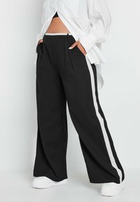 Czarne spodnie o szerokich nogawkach z białymi paskami po bokach, z plisami i płaskim pasem w talii, połączone z białymi butami i luźną białą koszulą.