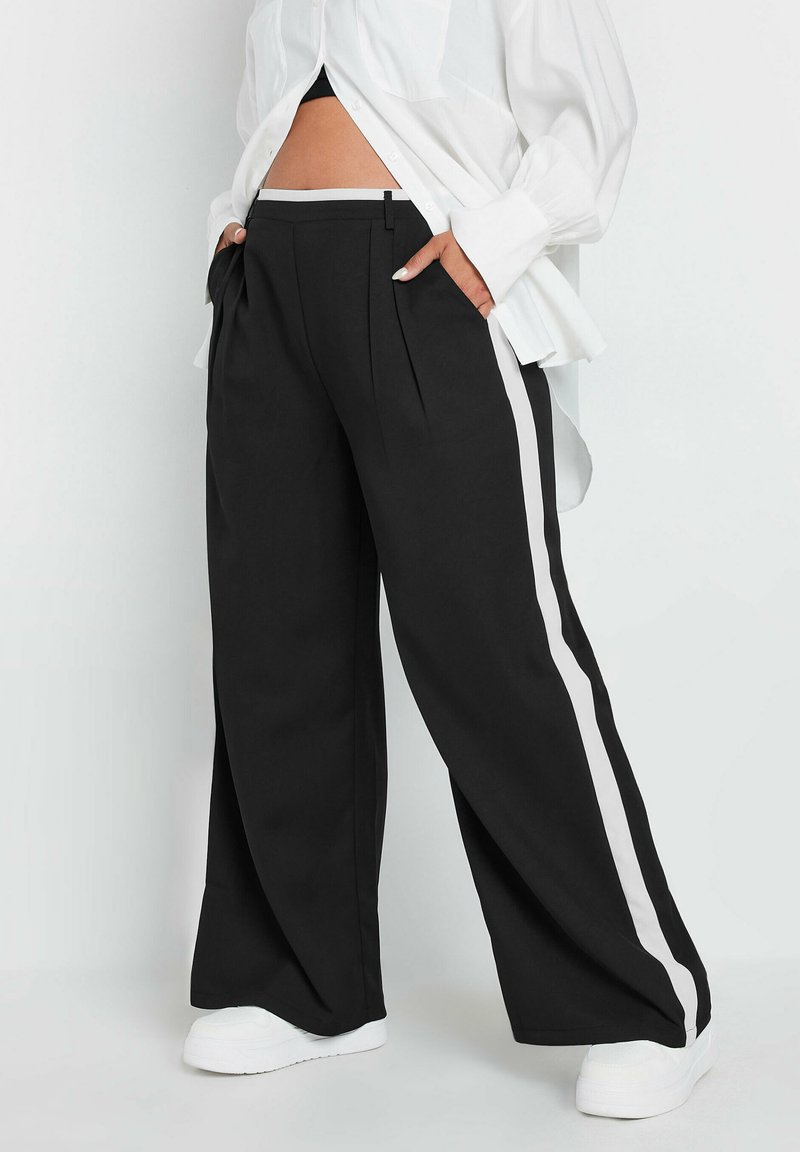 Czarne spodnie o szerokich nogawkach z białymi paskami po bokach, z plisami i płaskim pasem w talii, połączone z białymi butami i luźną białą koszulą.