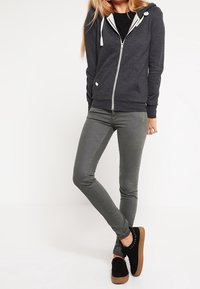 Tmavě šedá mikina na zip s bílými šňůrkami, pod ní černé tričko, doplněné úzkými šedými džínami a černými teniskami s hnědými podrážkami.