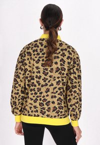 Transparent bomberjacka med leopardmönster i gult, svart och brunt. Har en gul ribbad krage och nederkant, med puffärmar.