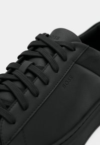 Κοντινή λήψη μαύρου δερμάτινου αθλητικού παπουτσιού με μαύρα κορδόνια, ανάγλυφο λογότυπο "BOSS" στο πλάι και στη γλώσσα, με ελάχιστες ραφές.