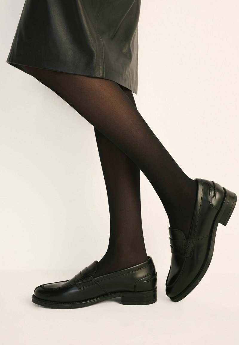 Czarne skórzane mokasyny o gładkiej fakturze, zaokrąglonym nosku i niskim obcasie, zestawione z czarnymi, przejrzystymi rajstopami. Projekt charakteryzuje się subtelnym szwem.