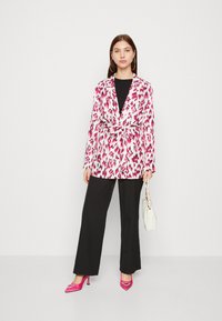 Blazer branco com estampado de animais em rosa, atado na cintura, combinado com calças largas pretas e sapatos de salto rosa, segurando uma bolsa branca.