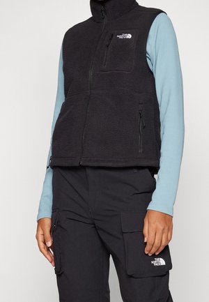 Personen har en sort fleecevest med lynlåslommer og en lyseblå langærmet skjorte på, kombineret med sorte cargobukser med klaplommer.