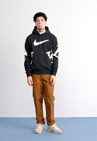 Μαύρη φούτερ με καπέλο της Nike με άσπρο λογότυπο και λεπτομέρειες, συνδυασμένη με καφέ παντελόνι. Ελαφριά αθλητικά παπούτσια σε ανοιχτό χρώμα. Ουδέτερο φόντο.